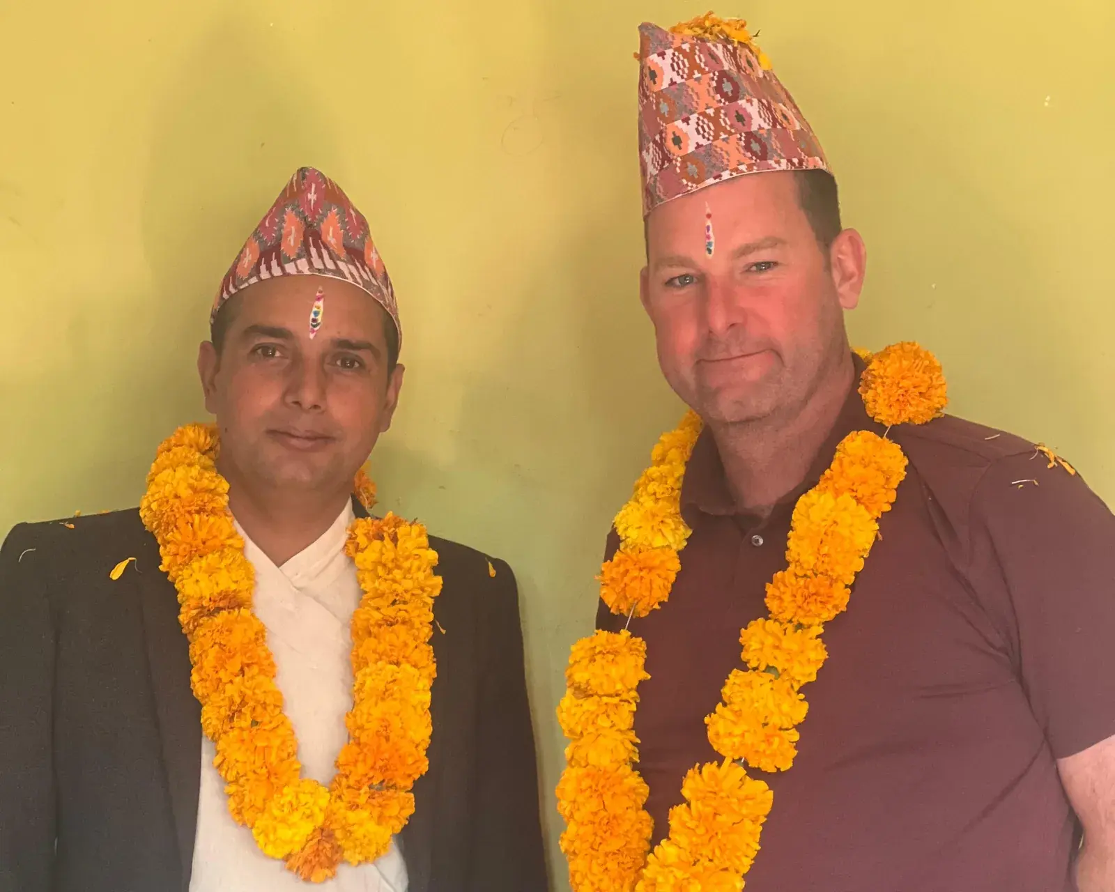 Joe and a Nepali friend side by side in topi caps and marigold garlands